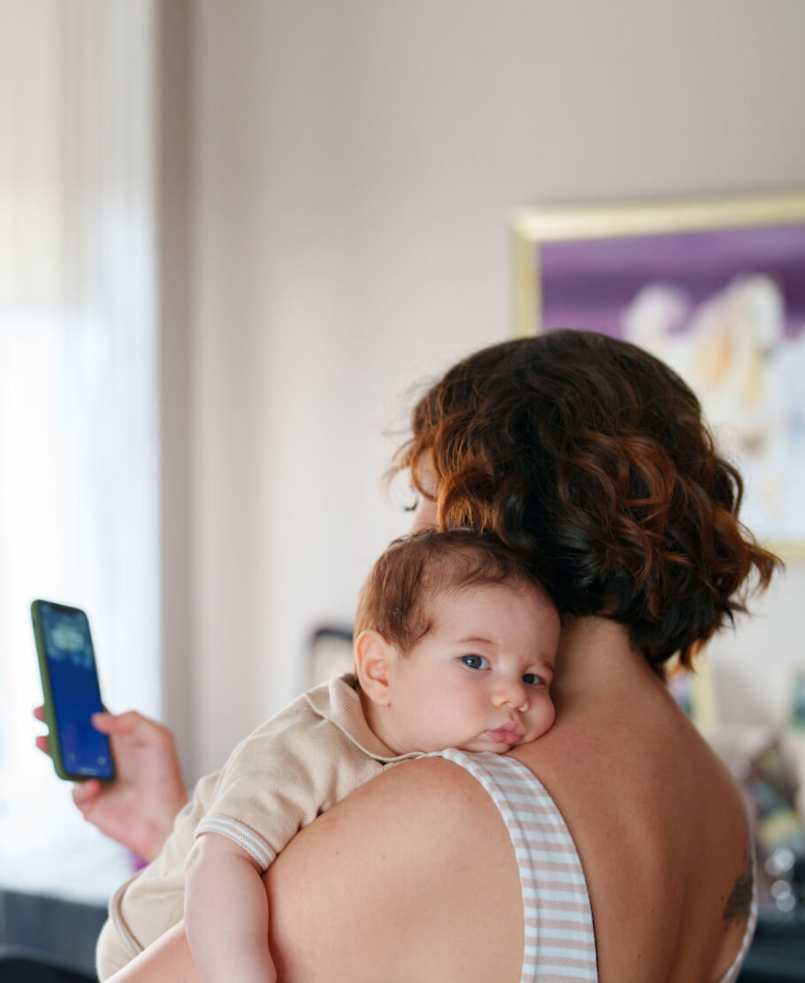 A mum facing away from the camera holding a baby over her shoulder and looking at her phone.
