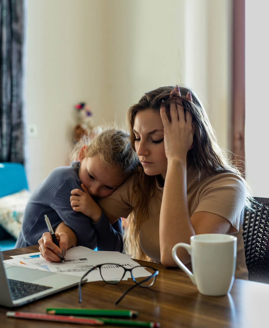 A mum sitting at a desk, writing on documents, while her toddler clings to her shoulder.
