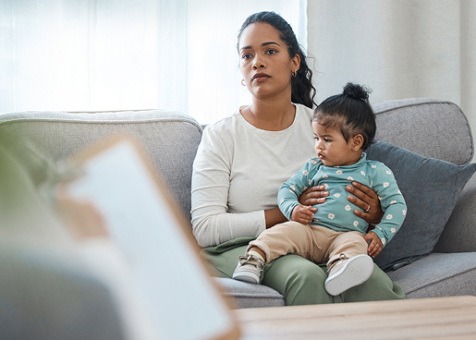 A mother sitting on a couch, holding her baby, facing the blurry figure of a person holding a clip-board.
